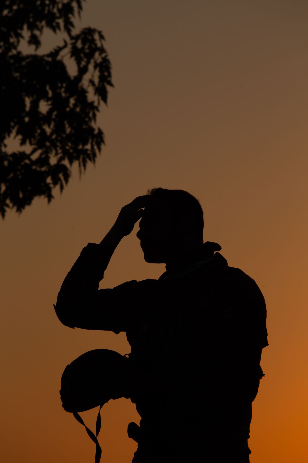 A veteran in silhouette holds his helmet, pausing in reflection against a warm sunset sky