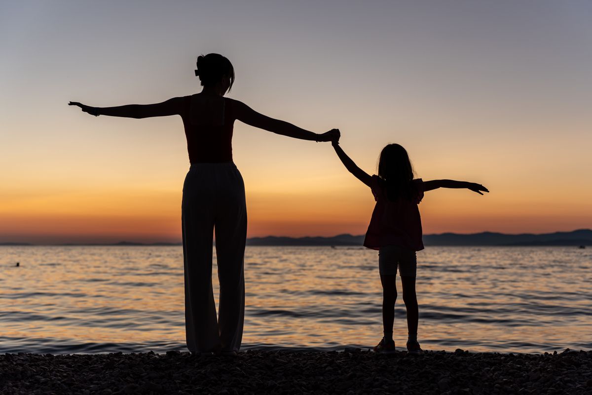 A mother and daughter stand in silhouette at the water's edge, arms raised together at sunset