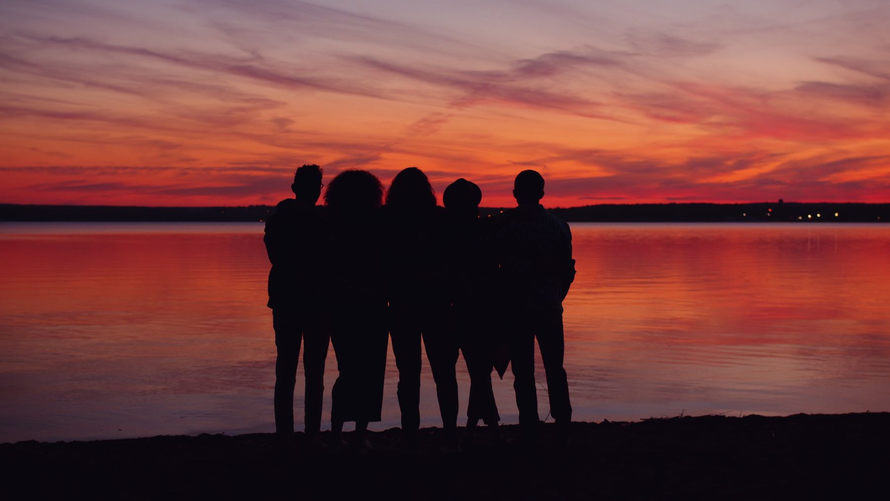 A Family stands together in silhouette on the shore at sunset, the sky glowing warm gold behind them