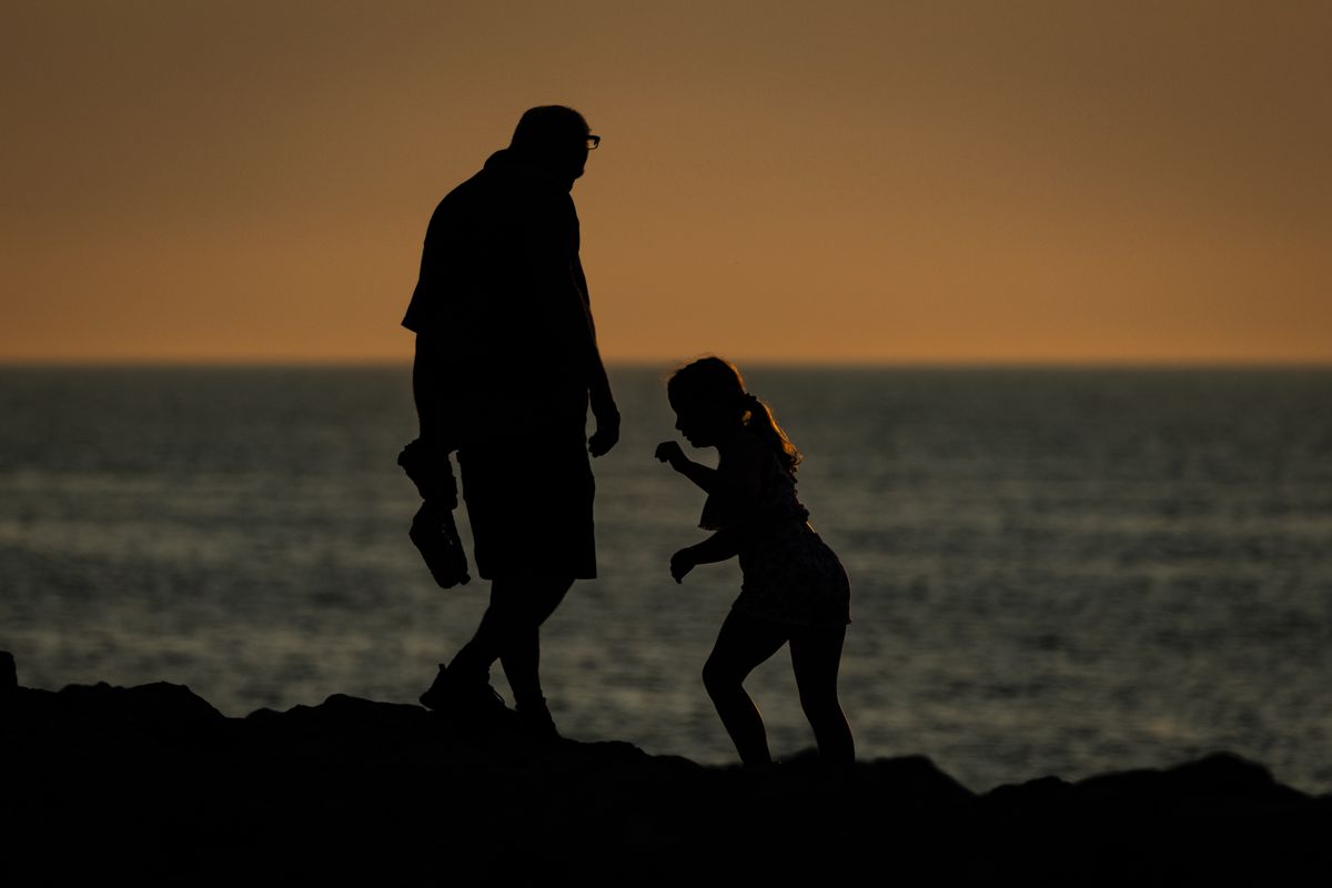 A father and his young daughter walk together in silhouette along a rocky shoreline at sunset, the ocean glowing behind them